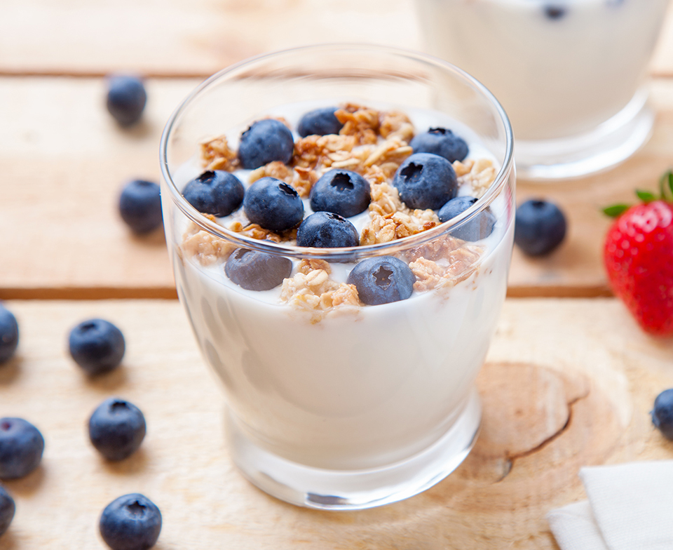 Glass cup of yogurt topped with granola and blueberries surrounded by loose berries.